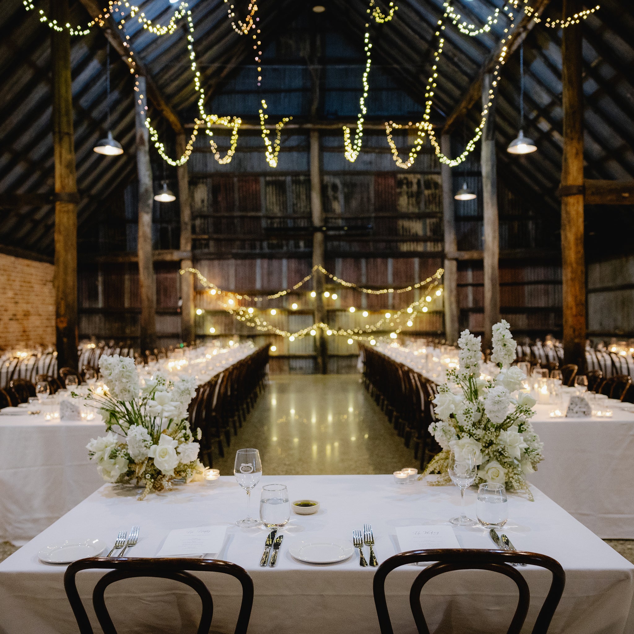 Rustic barn event setup with long white-linen tables, ambient candles, and wooden chairs — styled with Insieme Homewares terrazzo centrepieces and table numbers for an elegant yet relaxed celebration.