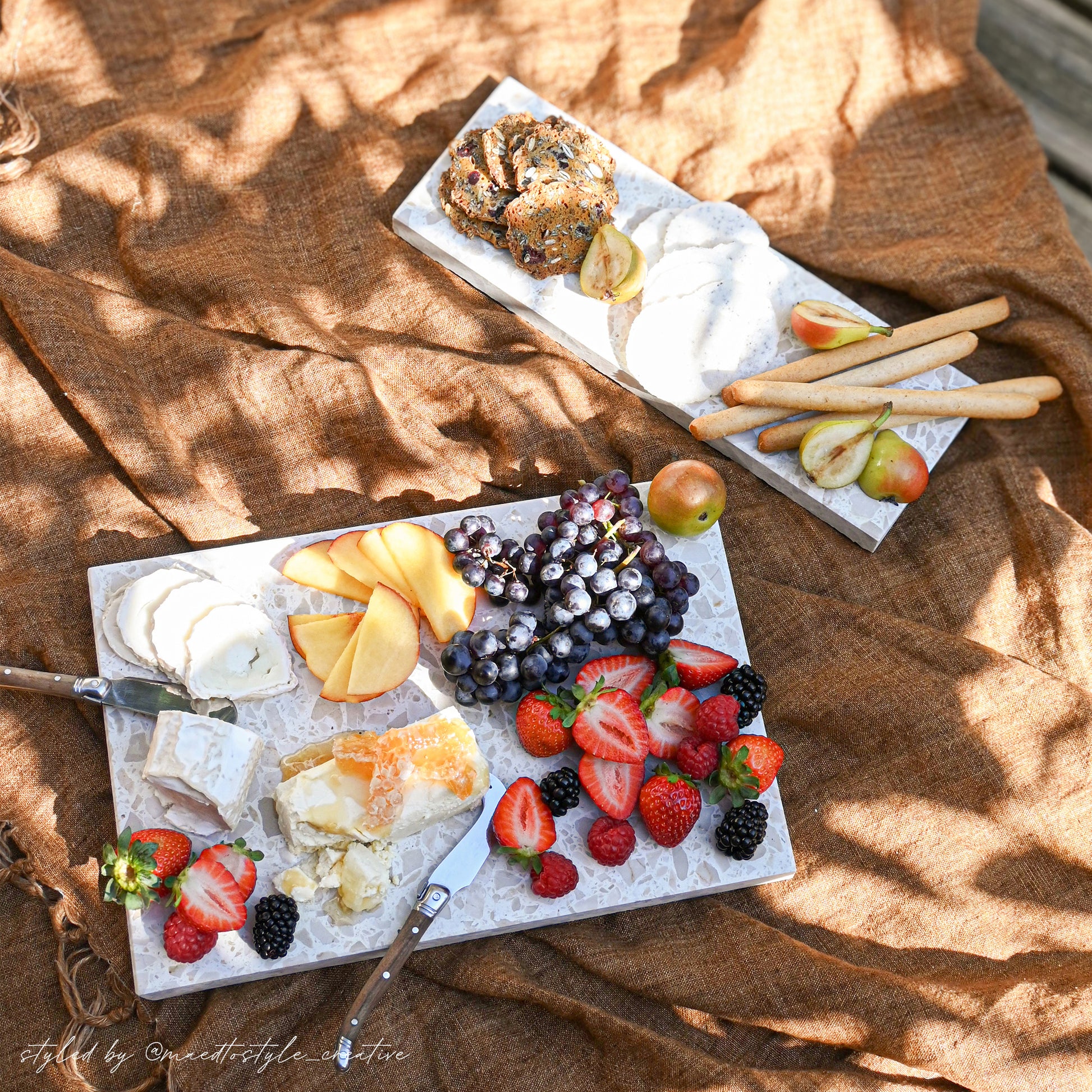 Insieme Homewares terrazzo platters styled outdoors with seasonal fruits, cheeses, and crackers — a sun-drenched picnic grazing setup on textured linen showcasing handcrafted stone serveware for alfresco entertaining.
