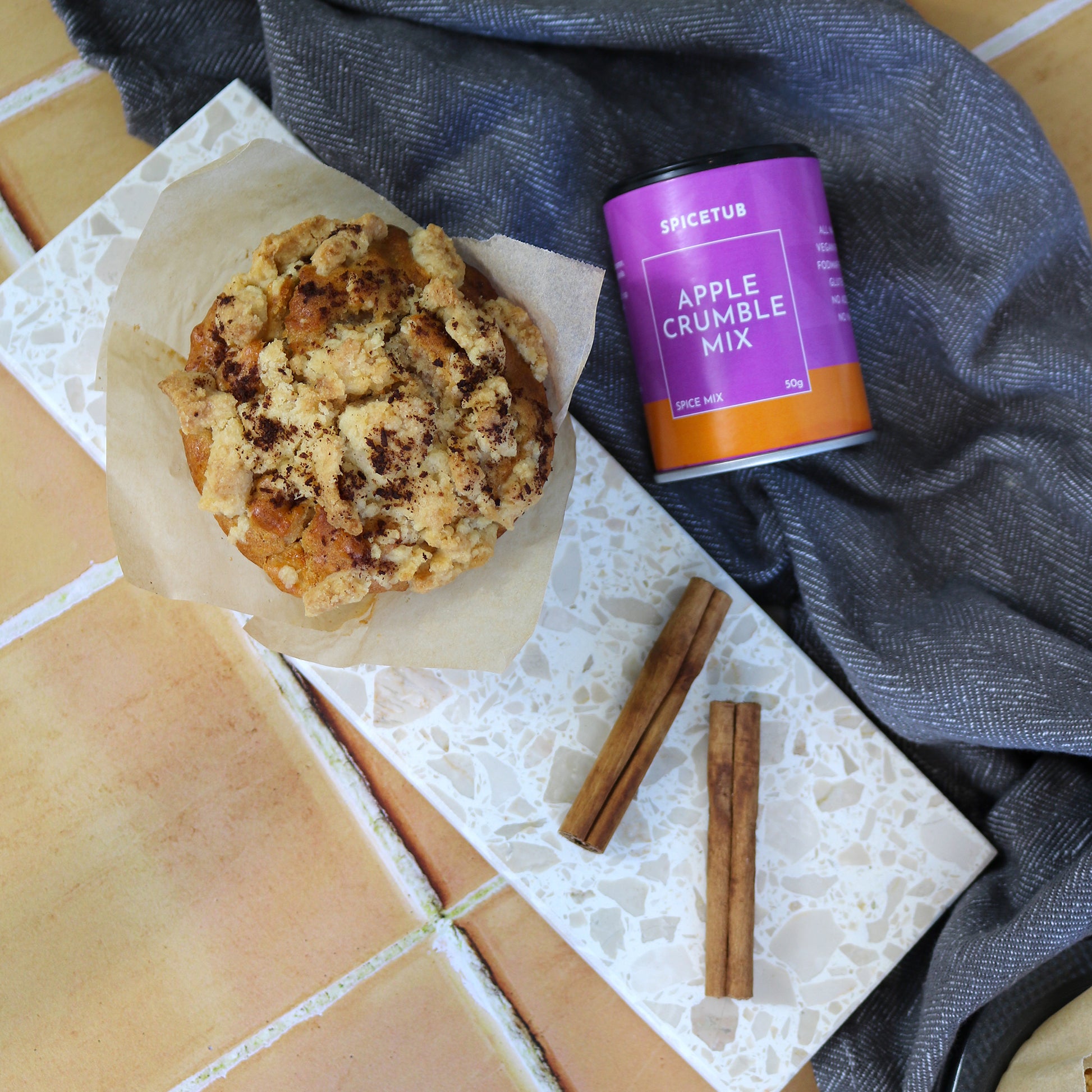 Flat lay of Apple Crumble Mix, cinnamon sticks, and baked goods atop a terrazzo platter, styled to evoke cosy, seasonal cooking.