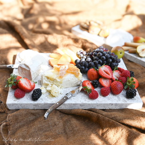 Insieme Homewares terrazzo platters styled outdoors with seasonal fruits, cheeses, and crackers — a sun-drenched picnic grazing setup on textured linen showcasing handcrafted stone serveware for alfresco entertaining.
