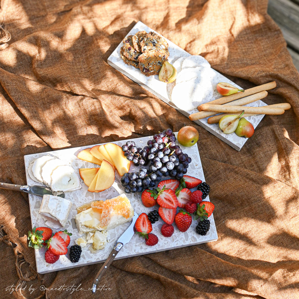 Insieme Homewares terrazzo platters styled outdoors with seasonal fruits, cheeses, and crackers — a sun-drenched picnic grazing setup on textured linen showcasing handcrafted stone serveware for alfresco entertaining.
