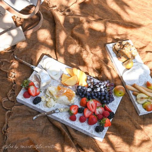 Insieme Homewares terrazzo platters styled outdoors with seasonal fruits, cheeses, and crackers — a sun-drenched picnic grazing setup on textured linen showcasing handcrafted stone serveware for alfresco entertaining.